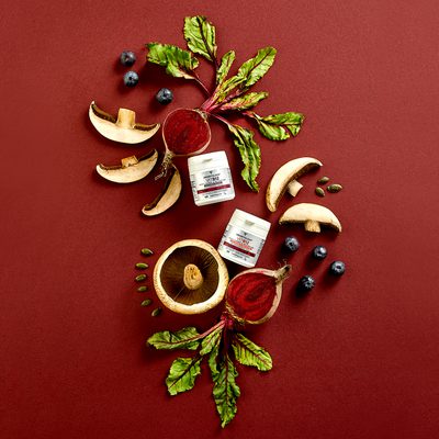 Two Vegetology Vit B12 tubs on a deep red background, arranged with beetroot, mushrooms, blueberries and pumpkin seeds