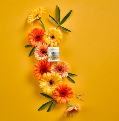 A Vegetology Vit D3 2500iu tub on a yellow background framed by orange and yellow gerberas