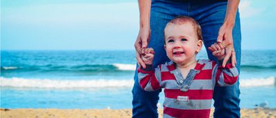 A smiling toddler holding an adult's hands on a beach