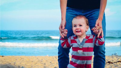 A smiling toddler holding an adult's hands on a beach